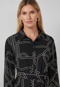 Zwarte jurk met contrasterende witte geometrische lijnen, kraag, lange mouwen, knoopsluiting aan de voorkant en een strikceintuur. Glad textuur.