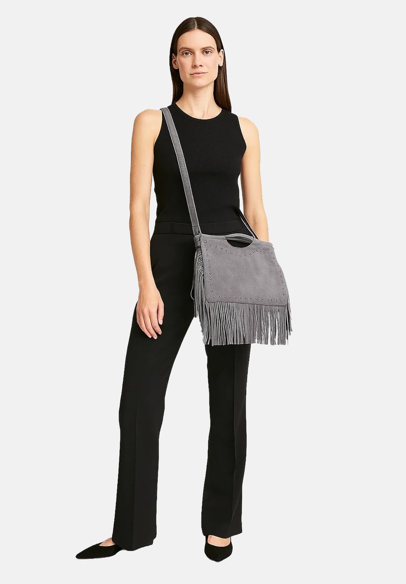 Sac en cuir suédé gris en bandoulière avec des franges, des clous décoratifs et une sangle, tenu par un modèle portant une tenue noire.