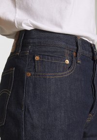 Pantalones vaqueros de mezclilla en azul oscuro con costuras claras. Presentan bolsillos frontales y acentos de hardware metálico. El material tiene una textura suave.