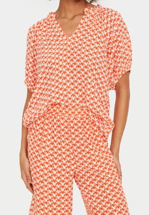 Femme portant un haut et un pantalon amples assortis avec un motif abstrait orange et blanc sur fond blanc.