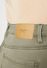 Gros plan sur la taille d'un pantalon en denim vert avec une étiquette en cuir beige portant l'inscription "VERO MODA DENIM" et une partie d'un haut côtelé beige.