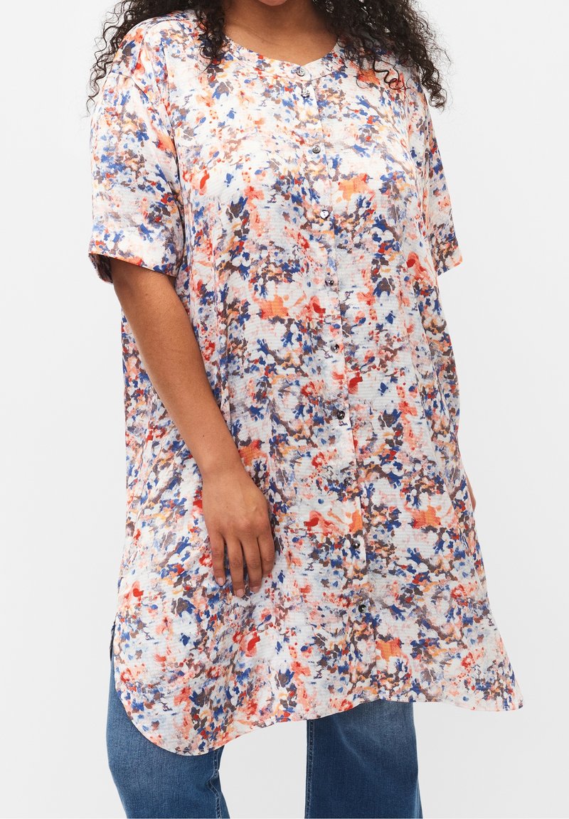 Chemise ample à manches courtes avec boutonnières, à motif abstrait multicolore, portée sur un jean bleu, visage du mannequin non visible.