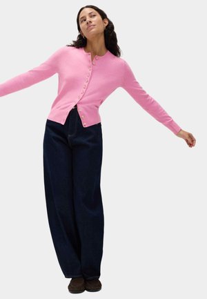 Cardigan rosa con scollo rotondo, bottoni frontali e maniche lunghe, abbinato a pantaloni di jeans scuri a vita alta e scarpe marrone scuro.