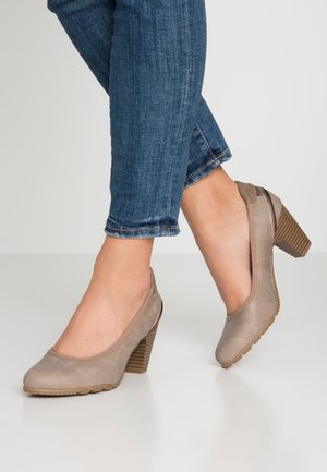 Kvinde iført beige ruskind blokhæl sko kombineret med opklemte blå jeans på en lysegrå gulv.