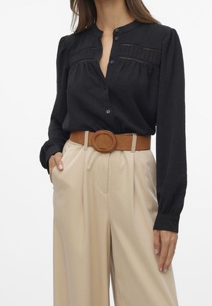 Kobieta w czarnej bluzce z długim rękawem z plisowanymi koronkowymi detalami oraz beżowych szerokich spodni z wysokim stanem, do których dodany jest pasek z okrągłą klamrą w kolorze brązowym.