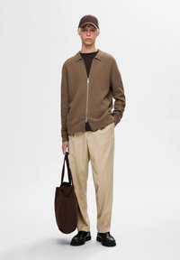 Cardigan marrón con cremallera sobre una camiseta oscura, pantalones de color beige y zapatos negros robustos. Sosteniendo un bolso tote marrón oscuro. Fit simple y relajado.