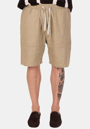 Pantaloncini di lino beige fino al ginocchio con cordoncino bianco, indossati con scarpe slip-on nere, che mostrano un tatuaggio geometrico nero sulla gamba destra.