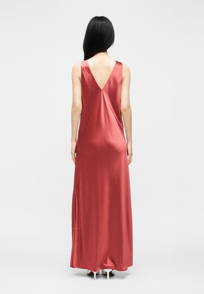 Γυναίκα με μακριά μαύρα μαλλιά στέκεται με την πλάτη προς το μέρος μας, φοράει ένα μακρύ ροζ-κόκκινο σατέν φόρεμα χωρίς μανίκια με V-κορδόνι στην πλάτη και κλείσιμο στο πλάι, και λευκά τακούνια.