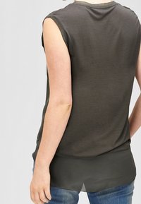 Oliven grøn ærmeløs top med glat tekstur, udskåret ryg og transparent nederste lag, parret med denim jeans.