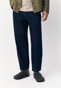 Pantalon en denim bleu foncé à coupe décontractée, jambes droites, avec des détails de coutures visibles. Associé à des chaussures en feutre gris et un haut beige.
