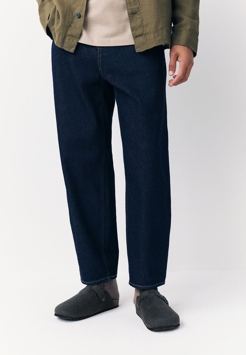 Pantalon en denim bleu foncé à coupe décontractée, jambes droites, avec des détails de coutures visibles. Associé à des chaussures en feutre gris et un haut beige.