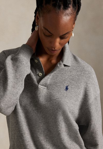 Woman with braided hair wearing a grey collared sweater with a small blue embroidered logo, touching her neck and looking downward.