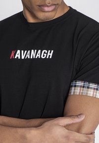 Camiseta negra con "KAVANAGH" en texto blanco y rojo, que presenta un puño con patrón de cuadros en beige, navy y blanco. Diseño suave y ajustado.