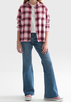Rød og pink ternet flannelsskjorte med knaplukning, båret over en hvid t-shirt. Kombineret med lyseblå, vide jeans og pink sneakers.