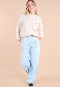 Un pull en tricot beige avec un motif texturé, associé à un jean large bleu clair ornés de broderies et de graphiques. Des baskets noires complètent la tenue.