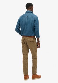 Mand set bagfra iført en blå denimskjorte, brune op-rullede bukser, brun læderbælte og lysebrune læderstøvler mod en hvid baggrund.