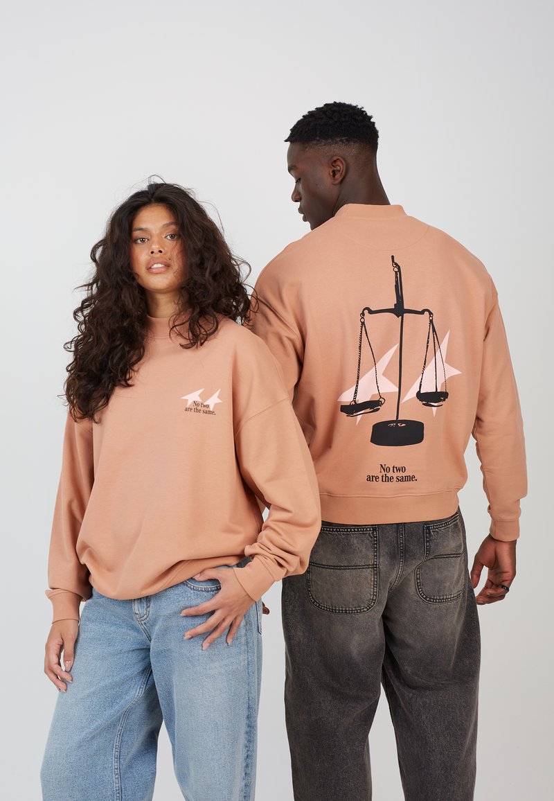 Light brown oversized sweatshirt featuring a graphic of a scale on the back and a small logo on the front, paired with loose denim jeans.