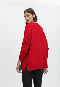 Roter Pullover mit langen Ärmeln, gerippten Bündchen und seitlichen Schlitzen am Saum, aus einem weichen Stoff gefertigt, kombiniert mit einer schwarzen Hose.