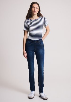 Femme debout portant un t-shirt à manches courtes blanc et bleu marine à rayures, un jean bleu foncé et des baskets blanches avec des rayures noires sur un fond uni.