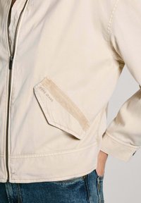 Beige bomullsjacka med dragkedja fram, en diagonal bröstficka med en mockadetalj, och ribbade ärmslut. Tyget har en mjuk textur.