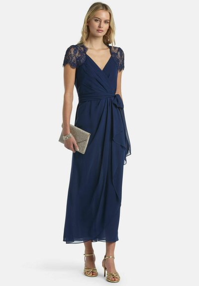 Femeie purtând o rochie înfășurată bleumarin cu mâneci scurte din dantelă, ținând o geantă plic strălucitoare și purtând sandale aurii cu toc înalt și bretele.