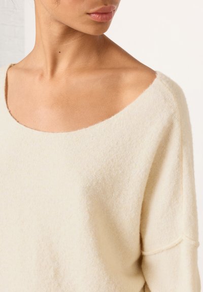 Femme portant un pull doux à large col de couleur crème, dévoilant son décolleté, sa clavicule et une partie de son visage tournée de profil.