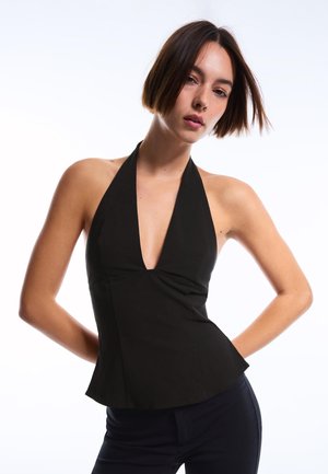 Mlada žena s kratkom smeđom kosom koja nosi crni top bez rukava s golim leđima i tamne hlače na bijeloj pozadini.