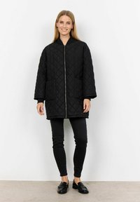 Svart vadderad jacka med blixtlås framtill, två framfickor och hög krage, kombinerad med svarta tajta byxor och loafers.