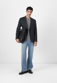 Schwarze strukturierte Blazer über einem grauen T-Shirt, kombiniert mit hellblauen, weiten Jeans und schwarzen Schuhen. Hält eine schwarze Laptoptasche.