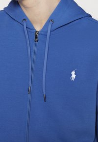 Blå zip-up hoodie i mjukt tyg med huva, med en vit polo-logotyp och svart metallblixtlås, tunna dragsnören och ribbade muddar.