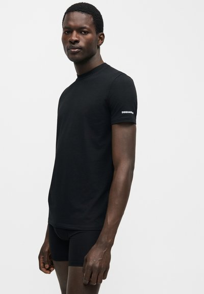 Άνδρας που στέκεται στο πλάι, φορώντας εφαρμοστή μαύρη μπλούζα με τη λέξη "DSQUARED2" στο μανίκι και μαύρα αντρικά εσώρουχα, σε απλό λευκό φόντο.