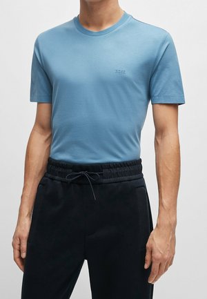 Mężczyzna ubrany w jasnoniebieski dopasowany t-shirt z logo "BOSS" i czarne spodnie ze sznurkiem w talii na jednolitym tle.