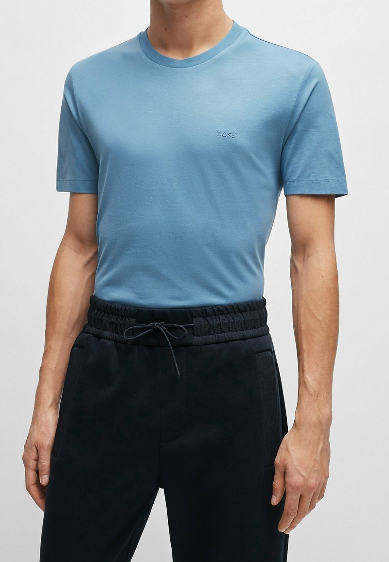 Homme portant un t-shirt bleu clair cintré avec le logo "BOSS" et un pantalon noir à cordon de serrage à la taille, sur un fond uni.
