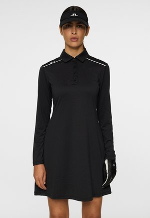 Frau in einem schwarzen langärmeligen Golfkleid, schwarzer Visiermütze und einem schwarz-weißen Golfhandschuh an der rechten Hand, vor einem schlichten Hintergrund stehend.