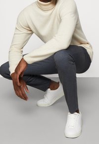 Maglione a collo alto in maglia beige, pantaloni slim-fit grigi a scacchi e sneakers bianche con punta arrotondata e lacci, posizionati su uno sfondo neutro.