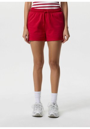 Kobieta w czerwonych szortach ze sznurkiem, białych skarpetkach do kostek i białych trampkach, stojąca z rozluźnionymi rękami wzdłuż ciała na białym tle.