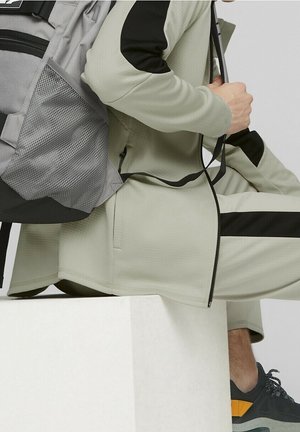 Personne portant un survêtement gris clair avec des accents noirs, assise sur un bloc blanc, ajustant la sangle d'un grand sac à dos gris, portant des baskets noires et orange.