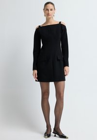 Mustat olkaimeton mekko pitkillä hihoilla, muotoiltu malli, kaksi etutaskua ja sileä pinta. Mallilla on päällä läpinäkyvät sukkahousut ja mustat kengät.