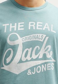 Světle modré bavlněné tričko s bílým grafickým textem "THE REAL JACK & JONES" s vintage stylem loga a hladkou strukturou.