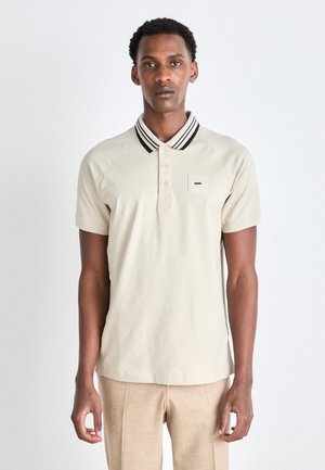 Beige polo shirt with black and white striped collar, short sleeves, and a small logo patch on the left chest. Clean design and soft texture.