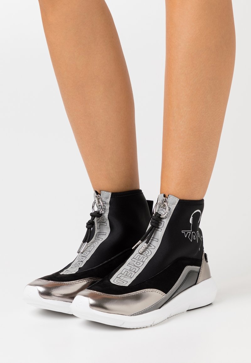 Czarne i metalicznie srebrne sneakersy za kostkę z grubymi białymi podeszwami, zamek z przodu i logo "Karl Lagerfeld", noszone na gołych nogach.