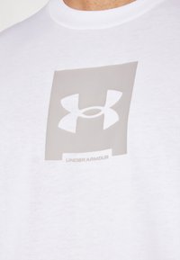 Hvid bomulds t-shirt med et stort gråt Under Armour-logo i midten, med en glat tekstur og en klassisk rund halsudskæring.