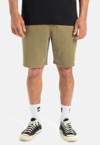 Olive green cotton shorts with a straight cut, slight stretch, and visible stitching details. Paired with black canvas sneakers and white socks.