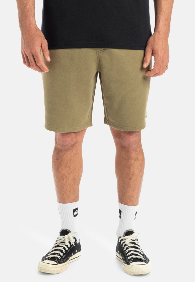 Olive green cotton shorts with a straight cut, slight stretch, and visible stitching details. Paired with black canvas sneakers and white socks.