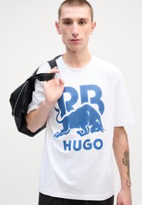 Hvid bomulds T-shirt med et stort blåt bøf grafik og "HUGO" tekst. Modellen holder en sort taske over skulderen.