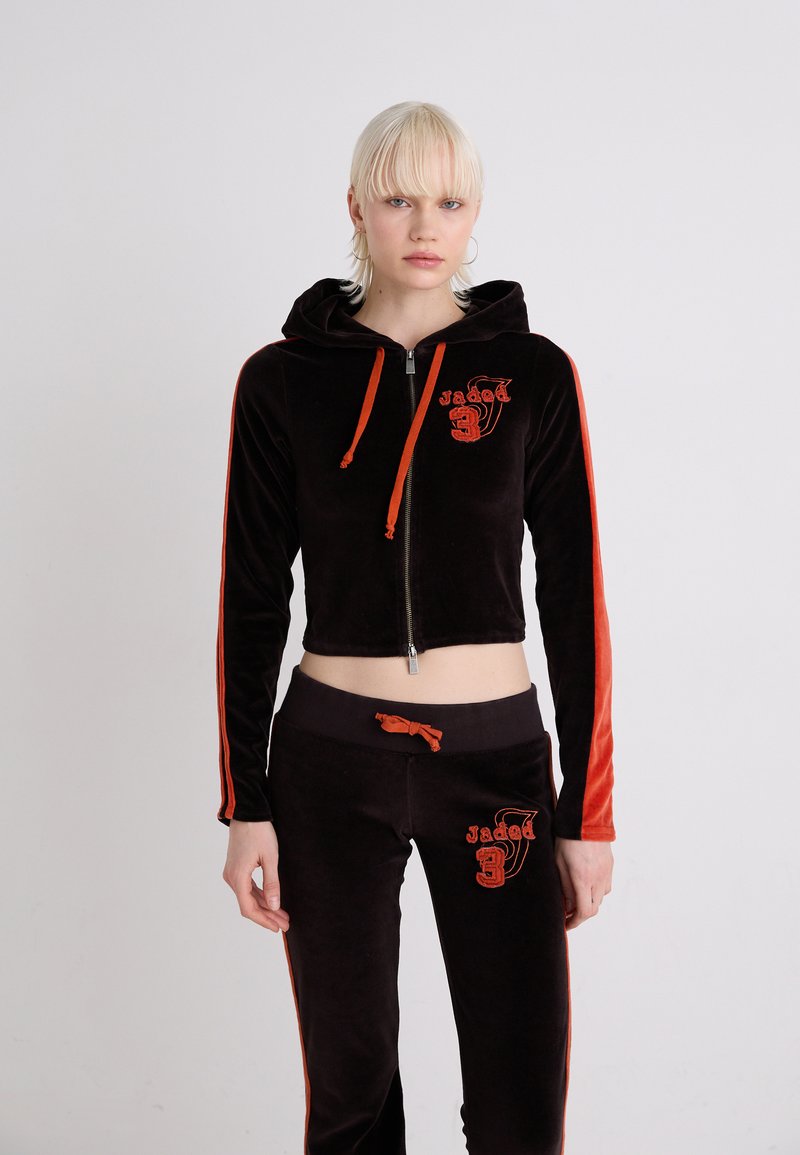 Felpa corta in velluto nero con accenti rossi, chiusura con zip e logo sul petto. Pantaloni neri abbinati con logo, vita elastica e cordino.