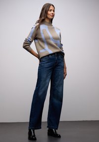 Sweter dzianinowy w kolorze beżowym z niebieskim abstrakcyjnym wzorem, zestawiony z szerokimi, ciemnoniebieskimi jeansami i czarnymi botkami za kostkę. Gładka faktura, luźny krój.