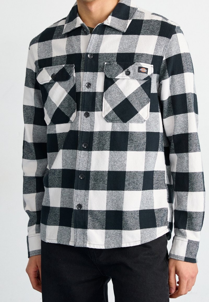 Homme portant une chemise en flanelle à carreaux noirs et blancs avec des poches poitrine boutonnées et un pantalon noir, torse et bras visibles.