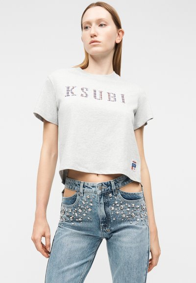 Γυναίκα που φοράει ανοιχτό γκρι κοντομάνικη μπλούζα με το κείμενο "KSUBI" και τζιν παντελόνι με ψηλή μέση σε μπλε χρώμα, διακοσμημένο με στρας και λεπτομέρειες με κοψίματα.