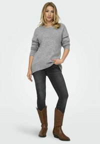 Suéter gris de punto con mangas largas y dobladillo acanalado; combinado con jeans oscuros y botas de cowboy marrones con un patrón acolchado.